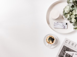 A minimalist scene featuring a white tray with a potted plant, a smartphone with a marble case, and a cylindrical container. Nearby, a cup of coffee sits beside an open magazine displaying images of architectural design.