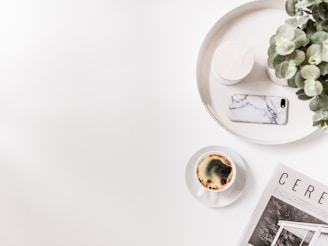 A minimalist scene featuring a white tray with a potted plant, a smartphone with a marble case, and a cylindrical container. Nearby, a cup of coffee sits beside an open magazine displaying images of architectural design.