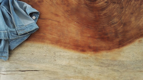 A denim jacket partially lying on a smooth, polished wooden surface. The wood has a distinct, swirling grain pattern with a mix of light and dark brown hues.