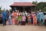 A large family group poses together in front of a traditional wooden house with a red roof. The group consists of adults, teenagers, and children, all wearing colorful traditional clothing typical of a cultural or festive occasion. The scene has a natural background with trees and parked cars.