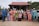 A large family group poses together in front of a traditional wooden house with a red roof. The group consists of adults, teenagers, and children, all wearing colorful traditional clothing typical of a cultural or festive occasion. The scene has a natural background with trees and parked cars.