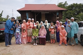 A large family group poses together in front of a traditional wooden house with a red roof. The group consists of adults, teenagers, and children, all wearing colorful traditional clothing typical of a cultural or festive occasion. The scene has a natural background with trees and parked cars.