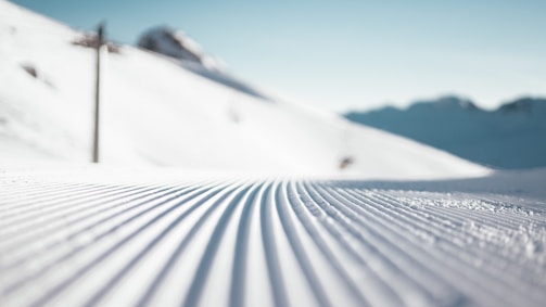 The image captures a close-up view of a well-groomed ski slope with finely combed snow patterns leading into the distance. In the background, snow-covered mountains are visible under a clear blue sky.