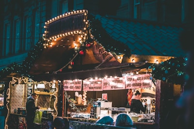 A cozy food stall adorned with festive lights and decorations, featuring a menu that includes various meats, potatoes, and sauces. People are gathered, ordering food and enjoying the warm atmosphere.