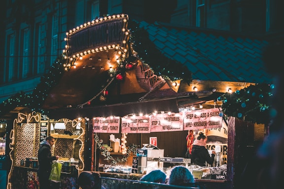 A cozy food stall adorned with festive lights and decorations, featuring a menu that includes various meats, potatoes, and sauces. People are gathered, ordering food and enjoying the warm atmosphere.