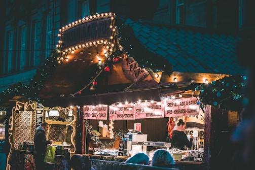 A cozy food stall adorned with festive lights and decorations, featuring a menu that includes various meats, potatoes, and sauces. People are gathered, ordering food and enjoying the warm atmosphere.