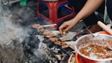 A hand is seen grilling skewers of meat over an outdoor charcoal stove surrounded by smoke and ash. Nearby, a bowl contains a spicy sauce with visible chili peppers. The setting appears to be informal, with plastic stools in the background.