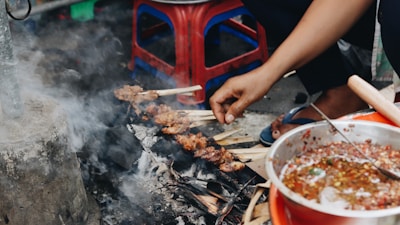 A hand is seen grilling skewers of meat over an outdoor charcoal stove surrounded by smoke and ash. Nearby, a bowl contains a spicy sauce with visible chili peppers. The setting appears to be informal, with plastic stools in the background.