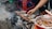 A hand is seen grilling skewers of meat over an outdoor charcoal stove surrounded by smoke and ash. Nearby, a bowl contains a spicy sauce with visible chili peppers. The setting appears to be informal, with plastic stools in the background.