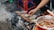 A hand is seen grilling skewers of meat over an outdoor charcoal stove surrounded by smoke and ash. Nearby, a bowl contains a spicy sauce with visible chili peppers. The setting appears to be informal, with plastic stools in the background.