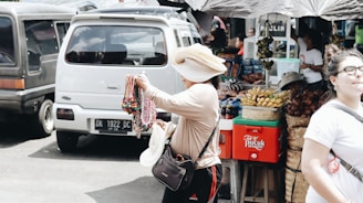 A street vendor is displaying an assortment of colorful necklaces and bracelets while wearing a large hat. Behind the vendor is a van and a food stall selling fruits and snacks, with people walking by.