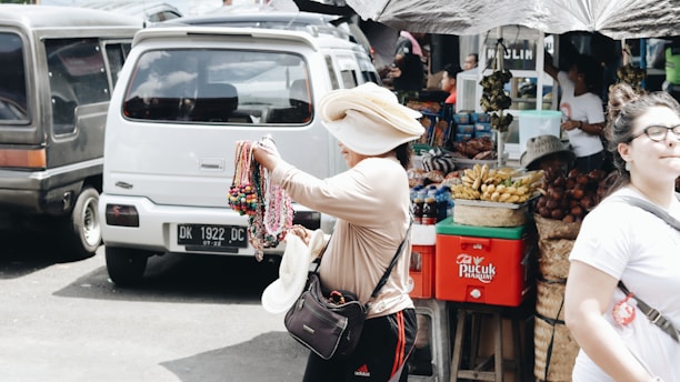 A street vendor is displaying an assortment of colorful necklaces and bracelets while wearing a large hat. Behind the vendor is a van and a food stall selling fruits and snacks, with people walking by.