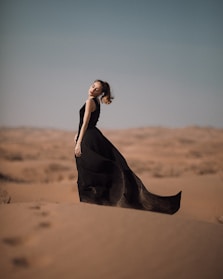 A graceful woman wearing a flowing black abaya with delicate embroidery, standing against a soft desert sunset.