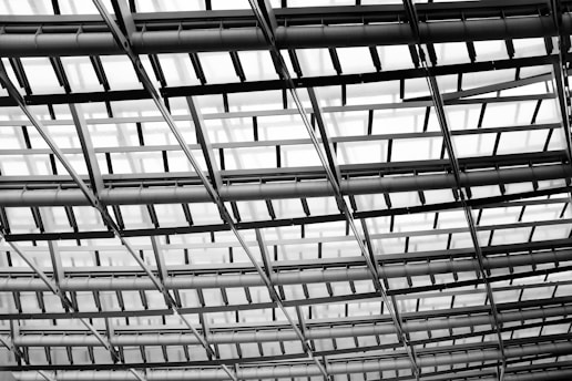 An intricate metal and glass structure creates a grid-like pattern with beams and panels, conveying a sense of modern architectural design.