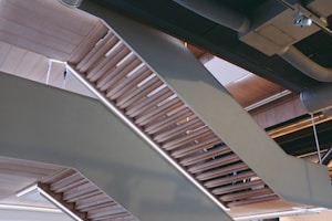An architectural structure featuring a series of modern, angular staircases with wooden steps and railings. Overhead, there are exposed pipes and ductwork, giving the interior a contemporary industrial look.