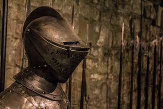 A medieval knight's armor is prominently displayed, focusing on the detailed helmet. The background features a stone wall and an array of long spears.
