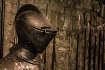 A medieval knight's armor is prominently displayed, focusing on the detailed helmet. The background features a stone wall and an array of long spears.