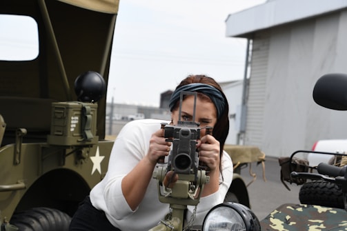 A person is aiming a vintage or military-style camera mounted on a vehicle equipped with camouflage patterns and a star symbol. The background shows an industrial setting with buildings and parked cars. The person appears to be concentrating while looking through the camera viewfinder.