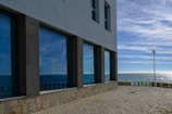 A large modern building with reflective windows faces the ocean. The reflection of the sky and the sea is visible in the windows, creating a vibrant play of blue shades. A lamppost stands near a stone-paved area that leads to the edge, overlooking the water. The sky above is clear with wispy clouds, and the sunlight sparkles on the surface of the sea.