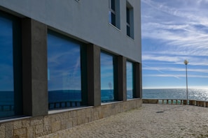 A large modern building with reflective windows faces the ocean. The reflection of the sky and the sea is visible in the windows, creating a vibrant play of blue shades. A lamppost stands near a stone-paved area that leads to the edge, overlooking the water. The sky above is clear with wispy clouds, and the sunlight sparkles on the surface of the sea.