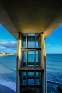 A modern elevator structure stands against the backdrop of an expansive ocean view. The blue waters of the sea stretch out under a partly cloudy sky, with the coast visible to the left. The architecture of the elevator features large glass panels framed by concrete, casting shadows along its length.