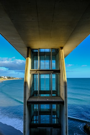 A modern elevator structure stands against the backdrop of an expansive ocean view. The blue waters of the sea stretch out under a partly cloudy sky, with the coast visible to the left. The architecture of the elevator features large glass panels framed by concrete, casting shadows along its length.
