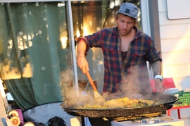 A person wearing a plaid shirt and cap is cooking outdoors using a large pan on a stove. Steam is rising from the dish being stirred with a wooden spatula. The setting appears to be a casual, sunlit outdoor area with reflections and greenery in the background.