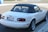 A white convertible sports car with a black soft top is parked on the side of a street. The vehicle has rounded taillights and a shiny finish. The license plate reads 'California'.