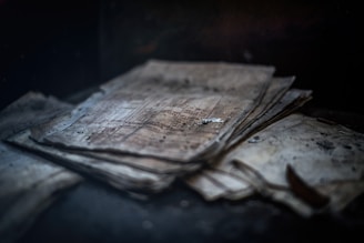 A stack of aged, weathered papers lies in a dark, dimly lit environment. The papers are yellowed and have visible spots and stains, suggestive of significant age and neglect.