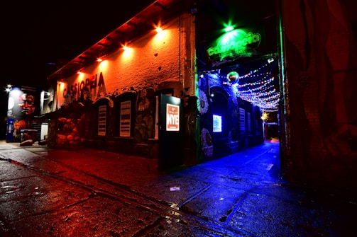 A vibrant nighttime street scene featuring a colorful building with the words 'Cassiopeia' glowing in red lights. The scene is adorned with a mix of neon lights and graffiti art. Festive string lights in blue, green, and red illuminate the alleyway, creating a lively atmosphere.