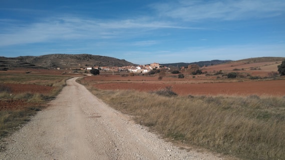 A rural landscape features a dirt road leading towards a small village in the distance. Surrounding the road are fields of dry grass and reddish-brown soil. The village consists of a cluster of buildings located at the base of a hilly terrain with scattered trees and vegetation. The sky above is clear with patches of clouds.