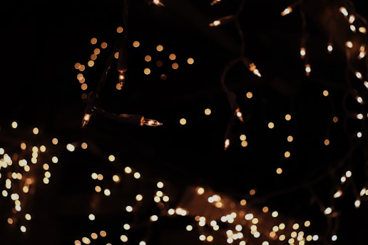 Strings of warm, glowing fairy lights create a soft, blurred bokeh effect against a dark background. The lights appear to hang down in an irregular pattern, casting a cozy and festive ambiance.