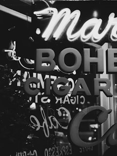 A nighttime city scene featuring illuminated neon signs in a combination of script and block letters. The word 'CIGAR' is prominently displayed alongside other partial words and reflections in the glass.