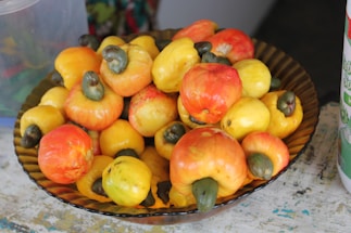 A decorative bowl filled with a vibrant assortment of ripe cashew apples, displaying shades of yellow, orange, and red. The cashew nuts, attached to the tops, are visible with their distinct dark green color.