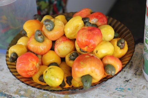 A decorative bowl filled with a vibrant assortment of ripe cashew apples, displaying shades of yellow, orange, and red. The cashew nuts, attached to the tops, are visible with their distinct dark green color.
