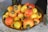 A decorative bowl filled with a vibrant assortment of ripe cashew apples, displaying shades of yellow, orange, and red. The cashew nuts, attached to the tops, are visible with their distinct dark green color.