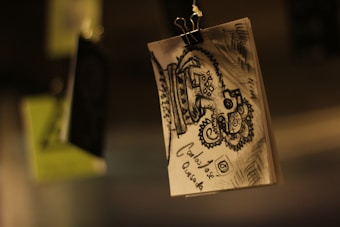 A sketch hangs from a clip, displaying an intricate black and white design featuring geometric patterns and swirling lines. The background is blurred, highlighting the paper on which the artwork is drawn. The lighting casts warm, muted tones over the scene.