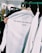 A close-up of a white shirt on a clothes hanger with the words 'OFF WHITE C/O VIRGIL ABLOH ©2013' printed on it. The shirt is being held by a person and is surrounded by other clothing items on hangers in a retail store environment. In the background, there are more clothes and a glimpse of a person using a phone.