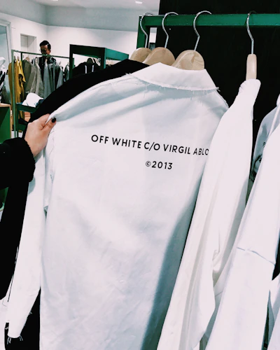 A close-up of a white shirt on a clothes hanger with the words 'OFF WHITE C/O VIRGIL ABLOH ©2013' printed on it. The shirt is being held by a person and is surrounded by other clothing items on hangers in a retail store environment. In the background, there are more clothes and a glimpse of a person using a phone.