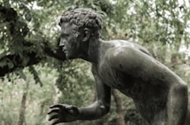 A bronze statue of a male figure in a dynamic, forward-leaning pose, set in a lush, green outdoor environment. The figure appears to be captured in motion, with a focused expression and detailed, curly hair.