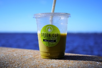 A transparent plastic cup with a green smoothie and a straw, placed on a ledge with a body of water and a clear blue sky in the background. The cup is labeled 'Green Cup Organic Cafe'.