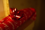 A close-up of a red gift box wrapped in red wrapping paper with white cursive writing. The box is adorned with a shiny red bow on top. The background is dimly lit, creating a warm and cozy atmosphere.