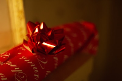 A close-up of a red gift box wrapped in red wrapping paper with white cursive writing. The box is adorned with a shiny red bow on top. The background is dimly lit, creating a warm and cozy atmosphere.