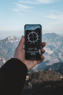A hand holding a smartphone displaying a compass app, with mountainous scenery in the background. The sky is clear with some clouds, and the compass on the phone screen shows a direction.