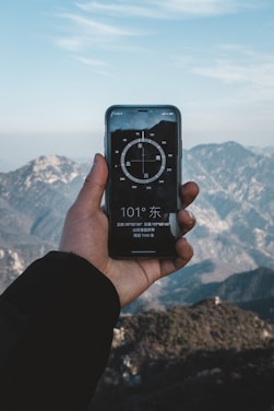 A hand holding a smartphone displaying a compass app, with mountainous scenery in the background. The sky is clear with some clouds, and the compass on the phone screen shows a direction.