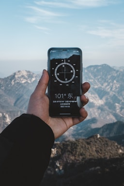 A hand holding a smartphone displaying a compass app, with mountainous scenery in the background. The sky is clear with some clouds, and the compass on the phone screen shows a direction.