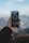 A hand holding a smartphone displaying a compass app, with mountainous scenery in the background. The sky is clear with some clouds, and the compass on the phone screen shows a direction.