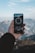 A hand holding a smartphone displaying a compass app, with mountainous scenery in the background. The sky is clear with some clouds, and the compass on the phone screen shows a direction.