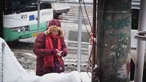 A person in a red parka with fur trim and a matching red scarf is standing on a snowy street, checking a smartphone. A bus with the number 413 is visible in the background, parked beside a snowy sidewalk. Snow is lightly falling, and a utility pole is in the foreground.