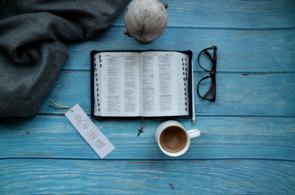 open book beside eyeglasses and mug